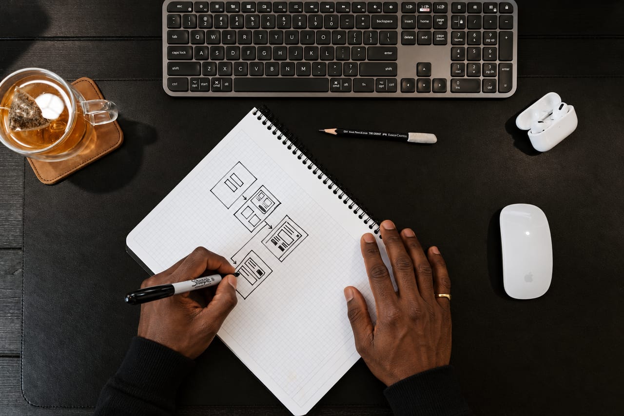 Hands sketching a wireframe flow of linked page layouts in a grid notebook, on a dark desk beside a keyboard.